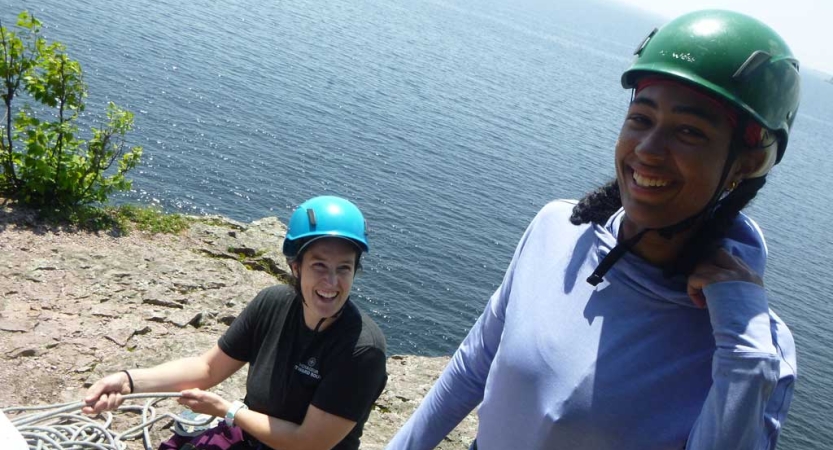 Two people wearing helmets smile at the camera. They appear to be at a high elevation. There is water below them.